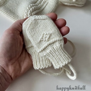 May include: A pair of white knitted baby booties with a matching white knitted hat. The hat has a tie string. The booties and hat are made with a textured knit pattern.