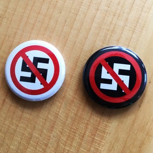 Puede incluir: Dos botones redondos con un círculo rojo y una línea diagonal que atraviesa una esvástica negra. Los botones están sobre una superficie de madera marrón claro.