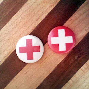 May include: Two red and white button pins with a white plus sign on a red background. The pins are on a wooden surface.