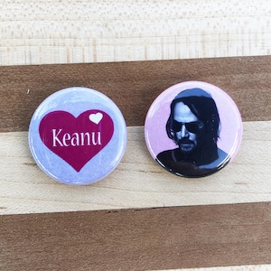 May include: Two round button badges. One badge has a purple background with a red heart that says "Keanu" in white text. The other badge has a pink background with a black and white image of a man with sunglasses.