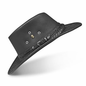 Op de afbeelding: Een zwarte leren cowboyhoed met een gevlochten band en een metalen gesp. De hoed heeft een decoratief reli&euml;fpatroon op de rand en een klein metalen leeuwenkopembleem op de kroon.