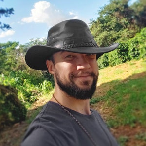 May include: A man with a beard wearing a black leather cowboy hat with a braided band. He is wearing a dark grey t-shirt and has a silver chain necklace around his neck. He is standing in front of a green grassy area with trees in the background.