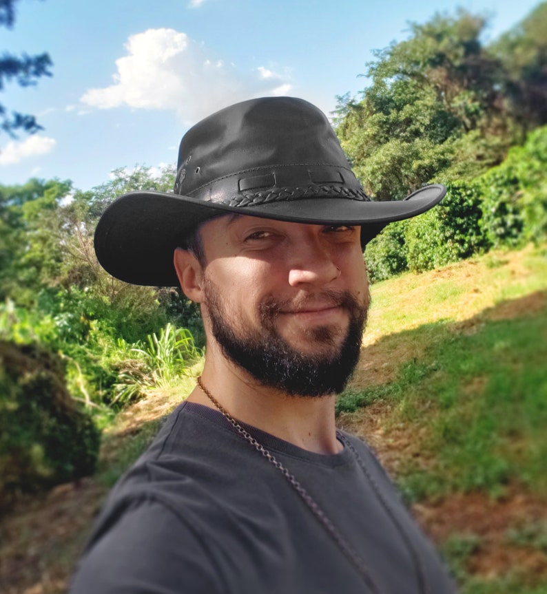 May include: A man with a beard wearing a black leather cowboy hat with a braided band. He is wearing a dark grey t-shirt and has a silver chain necklace around his neck. He is standing in front of a green grassy area with trees in the background.