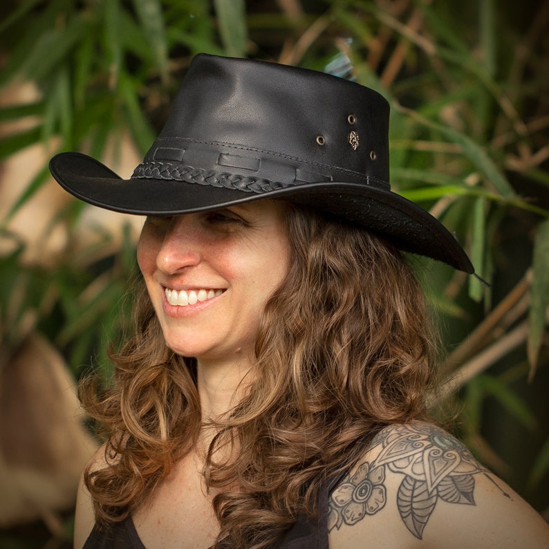 May include: A woman with curly brown hair is wearing a black leather cowboy hat with a braided band. She is smiling and looking to the side.
