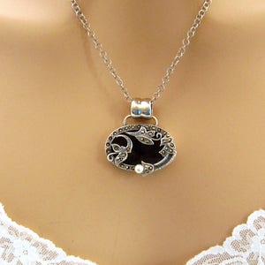 May include: A silver chain necklace with a black oval pendant featuring a floral design with marcasite stones and a pearl accent.
