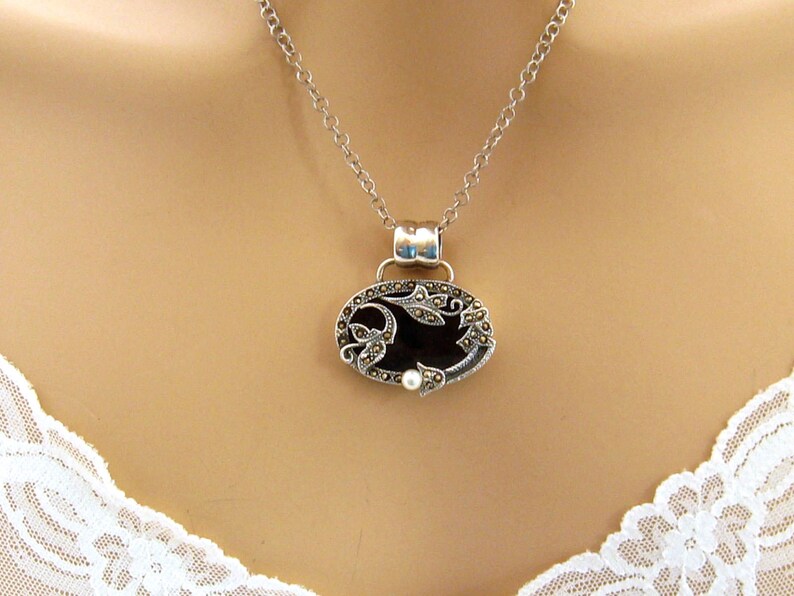 May include: A silver chain necklace with a black oval pendant featuring a floral design with marcasite stones and a pearl accent.
