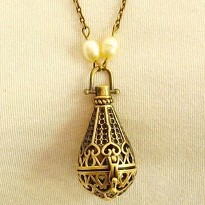 Op de afbeelding: Een gouden ketting met een delicate ketting en een hanger in de vorm van een traan. De hanger is ingewikkeld ontworpen met een filigraanpatroon en heeft een kleine opening aan de bovenkant. De ketting is versierd met twee witte parels.