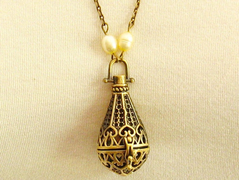 Op de afbeelding: Een gouden ketting met een delicate ketting en een hanger in de vorm van een traan. De hanger is ingewikkeld ontworpen met een filigraanpatroon en heeft een kleine opening aan de bovenkant. De ketting is versierd met twee witte parels.