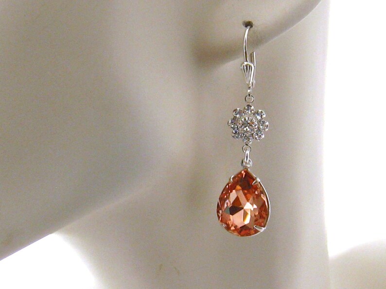 May include: A pair of silver dangle earrings with a teardrop-shaped peach-colored crystal and a smaller crystal cluster at the top.