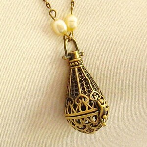 Op de afbeelding: Een gouden ketting met een delicate ketting en een hanger in de vorm van een traan. De hanger is gemaakt van ingewikkeld filigraanwerk en heeft een kleine opening aan de bovenkant. De ketting is versierd met kleine witte parels.