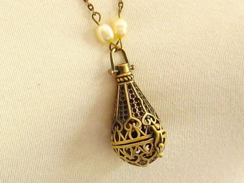 Op de afbeelding: Een gouden ketting met een delicate ketting en een hanger in de vorm van een traan. De hanger is gemaakt van ingewikkeld filigraanwerk en heeft een kleine opening aan de bovenkant. De ketting is versierd met kleine witte parels.