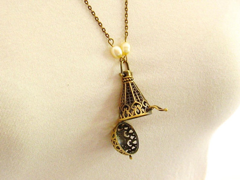 Op de afbeelding: Een gouden ketting met een filigraan hanger die opengaat om een klein, rond, opengewerkt compartiment te onthullen. De ketting is versierd met twee witte parels.