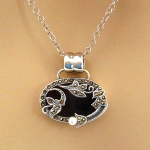 May include: A silver chain necklace with a black oval pendant featuring a floral design with a pearl accent.