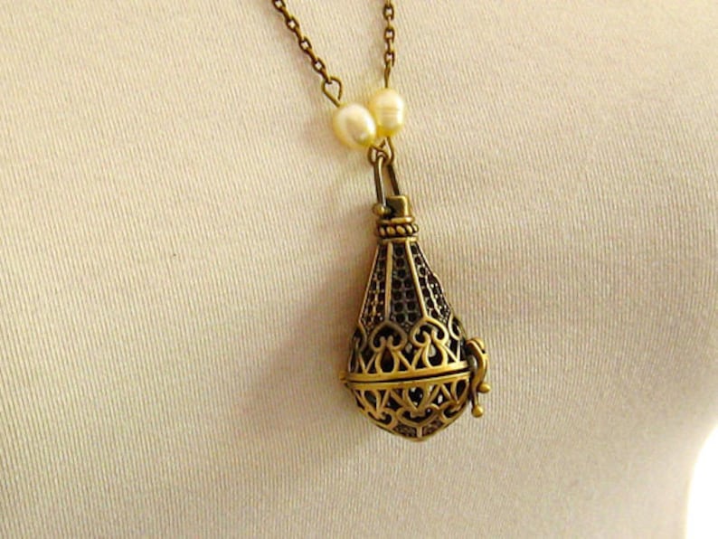 Op de afbeelding: Een gouden ketting met een delicate ketting en een hanger in de vorm van een kooi. De hanger heeft een filigraan ontwerp en is met een kleine lus aan de ketting bevestigd. Twee witte parels zijn aan de ketting boven de hanger bevestigd.