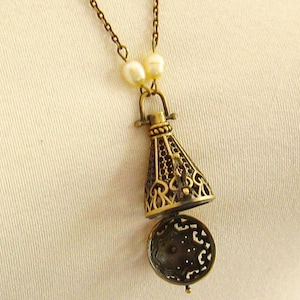 Op de afbeelding: Een gouden ketting met een decoratieve, opengewerkte hanger die lijkt op een klein, versierd flesje. De hanger is bevestigd aan een ketting met twee witte parel accenten.