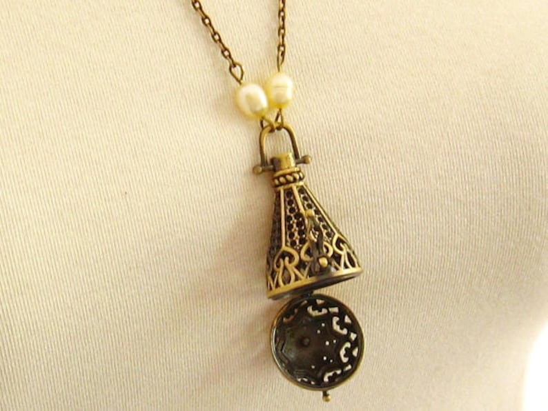 Op de afbeelding: Een gouden ketting met een decoratieve, opengewerkte hanger die lijkt op een klein, versierd flesje. De hanger is bevestigd aan een ketting met twee witte parel accenten.