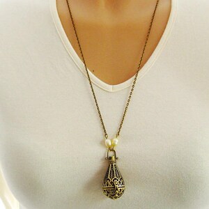 Op de afbeelding: Een gouden ketting met een decoratieve, filigraan hanger in de vorm van een traan. De hanger heeft een kleine opening aan de bovenkant, wat suggereert dat het een medaillon of een parfumflesje kan zijn.