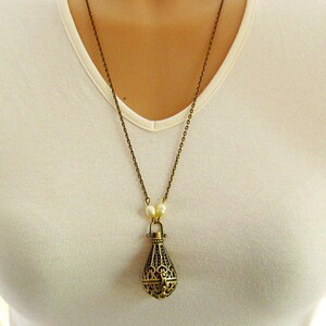 Op de afbeelding: Een gouden ketting met een kooivormige hanger en parel accenten. De hanger is traanvormig en heeft ingewikkelde details.