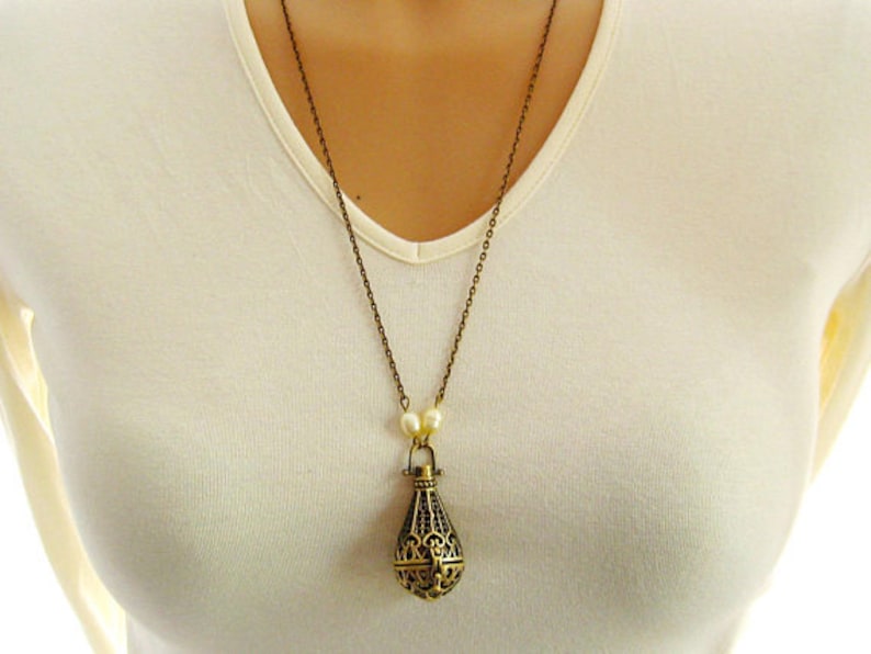 Op de afbeelding: Een gouden ketting met een kooivormige hanger en parel accenten. De hanger is traanvormig en heeft ingewikkelde details.