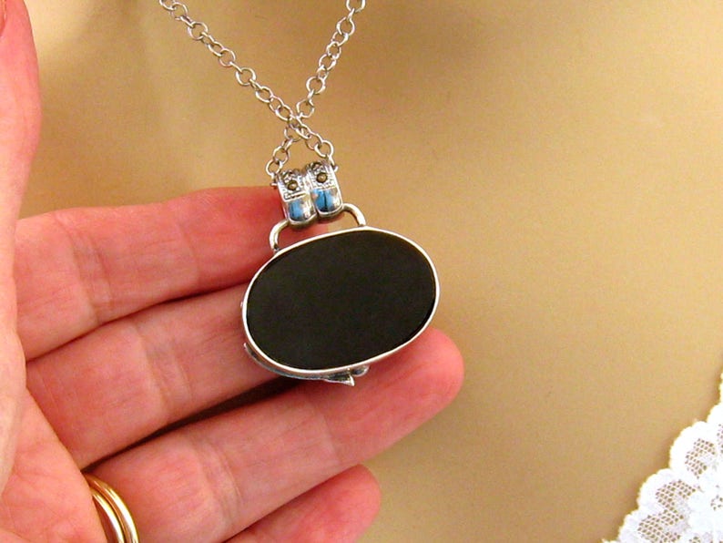 May include: A silver chain necklace with a black oval pendant. The pendant has a silver frame with blue accents.