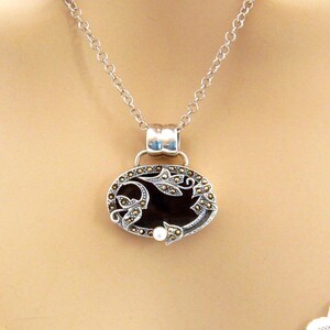 May include: A silver chain necklace with a pendant featuring a black oval cameo with a floral design and a single pearl accent.