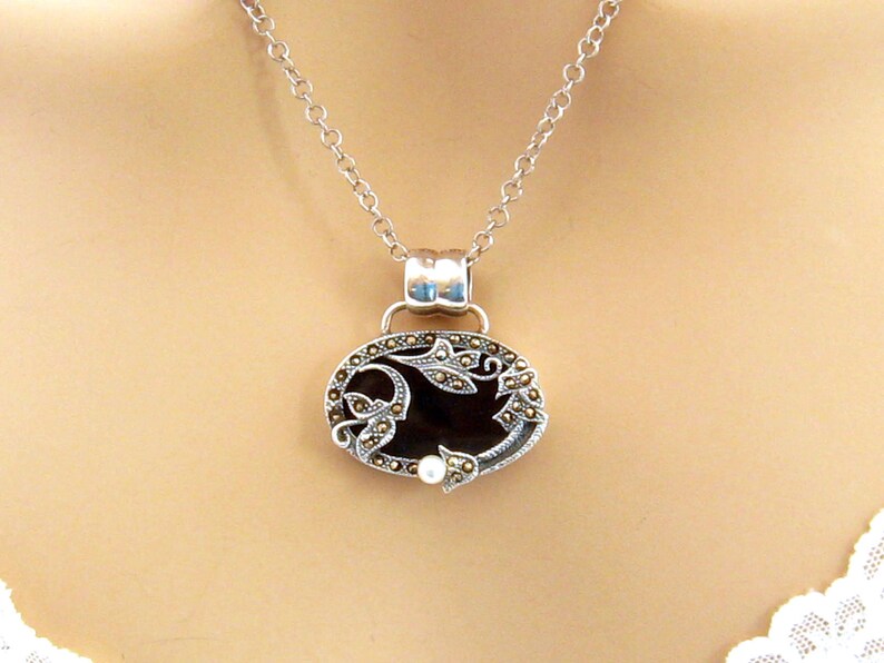 May include: A silver chain necklace with a pendant featuring a black oval cameo with a floral design and a single pearl accent.
