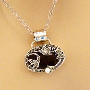 May include: A silver chain necklace with an oval pendant featuring a black onyx stone surrounded by a silver filigree design with marcasite accents. A small pearl is attached to the bottom of the pendant.
