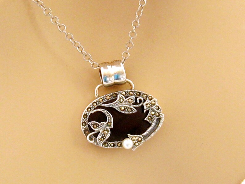 May include: A silver chain necklace with an oval pendant featuring a black onyx stone surrounded by a silver filigree design with marcasite accents. A small pearl is attached to the bottom of the pendant.