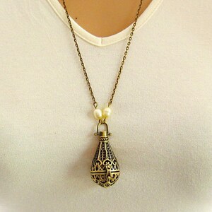 Op de afbeelding: Een gouden ketting met een delicate ketting en een hanger in de vorm van een kooi. De hanger is gemaakt van ingewikkeld metaalwerk en heeft een parel-achtige kraal aan de bovenkant.