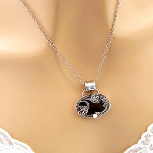 May include: A silver chain necklace with a black oval pendant featuring a floral design and a small pearl accent.