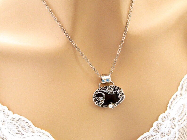 May include: A silver chain necklace with a black oval pendant featuring a floral design and a small pearl accent.