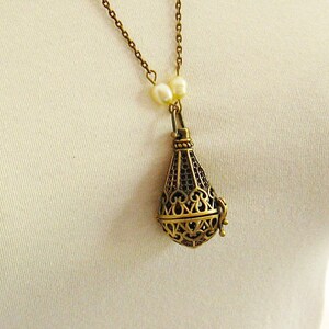 Op de afbeelding: Een gouden ketting met een delicate ketting en een hanger in de vorm van een kooi. De hanger is versierd met ingewikkelde ontwerpen en heeft een parel accent aan de bovenkant.