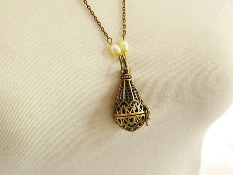 Op de afbeelding: Een gouden ketting met een delicate ketting en een hanger in de vorm van een kooi. De hanger is versierd met ingewikkelde ontwerpen en heeft een parel accent aan de bovenkant.