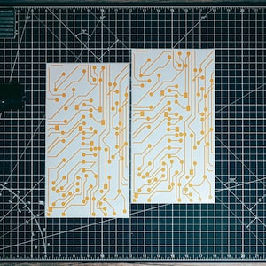 Könnte beinhalten: Zwei weiße, rechteckige Schablonen für Leiterplatten mit orangefarbenen Schaltungsdesigns werden auf einer schwarzen Schneidematte angezeigt. Eine amerikanische Flagge ist in die Matte geätzt. Eine schwarze Pistole und ein Bastelmesser sind ebenfalls sichtbar.