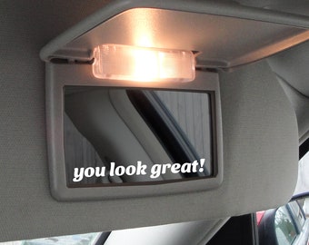 May include: A gray car visor with a built-in mirror and a light. The mirror has a white sticker that says "you look great!"