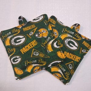 May include: Two square fabric pot holders featuring a dark green background with a repeating pattern of yellow and white Green Bay Packers logos, football helmets, pennants, and the text Green Bay Packers in a distressed yellow font.