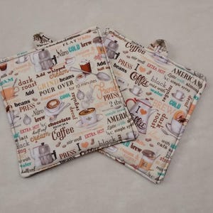 May include: Two cream-colored, square pot holders with a coffee-themed design. The fabric is covered with coffee-related words, coffee cups, and coffee-making equipment. Each pot holder has a small fabric loop for hanging.