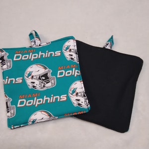 May include: Two square pot holders. One is teal with a repeating pattern of white and orange Miami Dolphins football helmets and the team name. The other is solid black. Both have a small fabric loop for hanging.