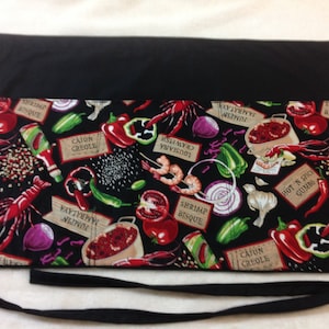 May include: Black waist apron with a colorful pattern of Cajun food items. The design includes red crawfish, peppers, tomatoes, and labels with text like "Cajun Creole" and "Hot 'N Spicy Gumbo". The apron has black ties.