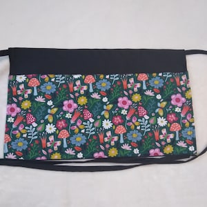 May include: A black waist apron with a colorful floral and mushroom pattern. The apron features a dark green background with pink, yellow, blue, and white flowers, butterflies, and red-capped mushrooms. The top band and ties are solid black.