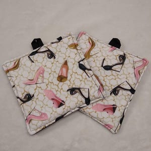 May include: Two square pot holders with a white background and a repeating pattern of high-heeled shoes in black, pink, and gold. Each pot holder has a black loop for hanging.