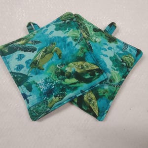 May include: Two square potholders with a loop for hanging. The fabric features a vibrant underwater scene with sea turtles, fish, and coral in shades of green, blue, and turquoise. The potholders are designed for kitchen use.