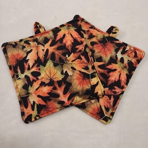 May include: Two square pot holders with a fall leaf pattern. The leaves are in shades of orange, yellow, and brown against a black background. Each pot holder has a small loop for hanging.