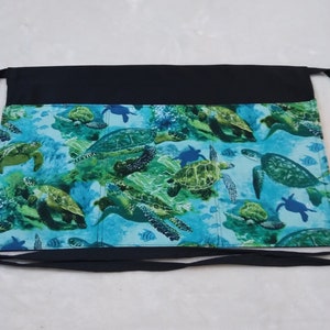 May include: Black and blue apron with a sea turtle design. The apron has two pockets and ties at the waist.