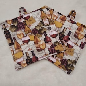 May include: Two square pot holders with a wine-themed pattern. The fabric features images of wine bottles, glasses of red wine, grapes, and cheese. The background is a cream color with French text. Each pot holder has a small fabric loop for hanging.