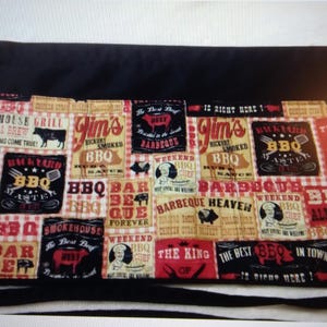 May include: Black apron with a patchwork design featuring BBQ-themed graphics in red, tan, and black. The apron includes text like "BBQ Master" and "Barbeque Heaven" and has a black tie.