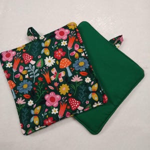 May include: Two square pot holders. One features a dark green background with a colorful floral pattern of butterflies, mushrooms, and flowers. The other is solid green. Both have a small fabric loop for hanging.