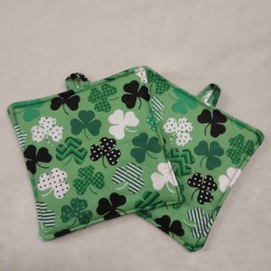 May include: Two square green pot holders with a St. Patrick's Day theme. The fabric features a pattern of shamrocks in various shades of green, black, and white. Each pot holder has a small loop for hanging.