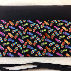 May include: Black waist apron with a colorful flip-flop pattern. The apron features a solid black top section and long black ties. The flip-flop design includes various colors like red, blue, green, and yellow, creating a vibrant, summery look.