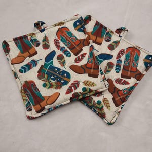 May include: Two square cream-colored pot holders with a repeating pattern of cowboy boots and feathers in shades of brown, teal, red, and blue. Each pot holder has a small fabric loop for hanging.